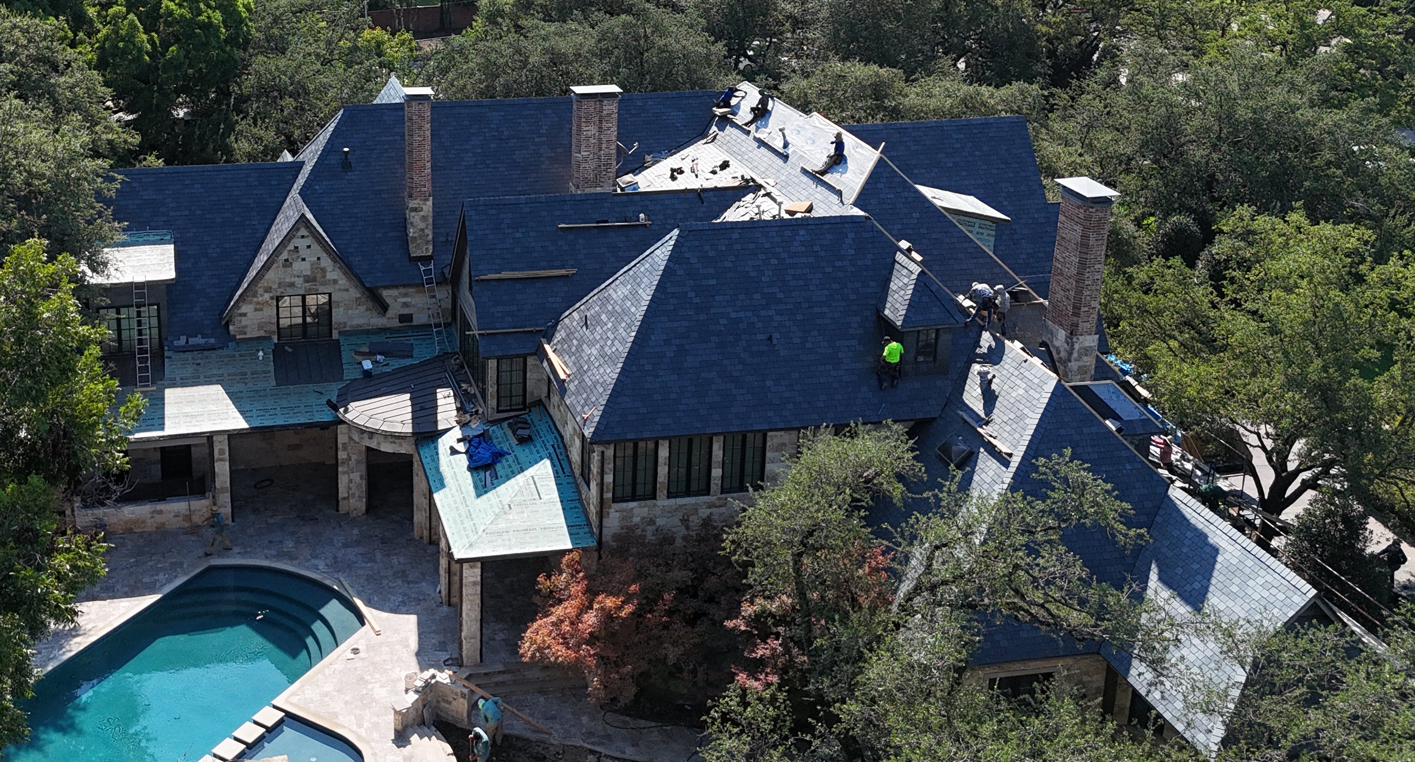 Drone view of a crew finishing a dark blue roof on a large stone home with a pool, surrounded by tall trees